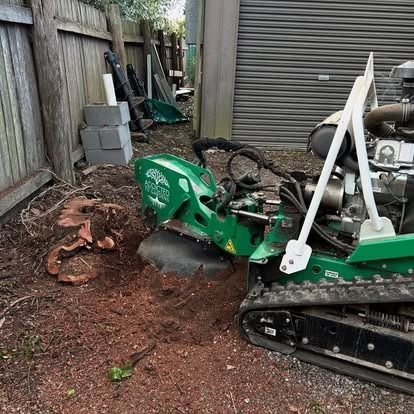 Stump grinder grinding a tree stump in a yard. Green machine, brown dirt, and gray garage door.
