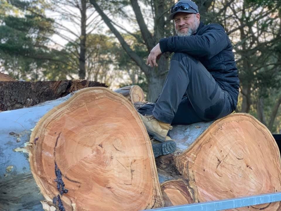 Man sitting on large, cut tree rounds. Outdoors, brown wood, black jacket, and gray pants.