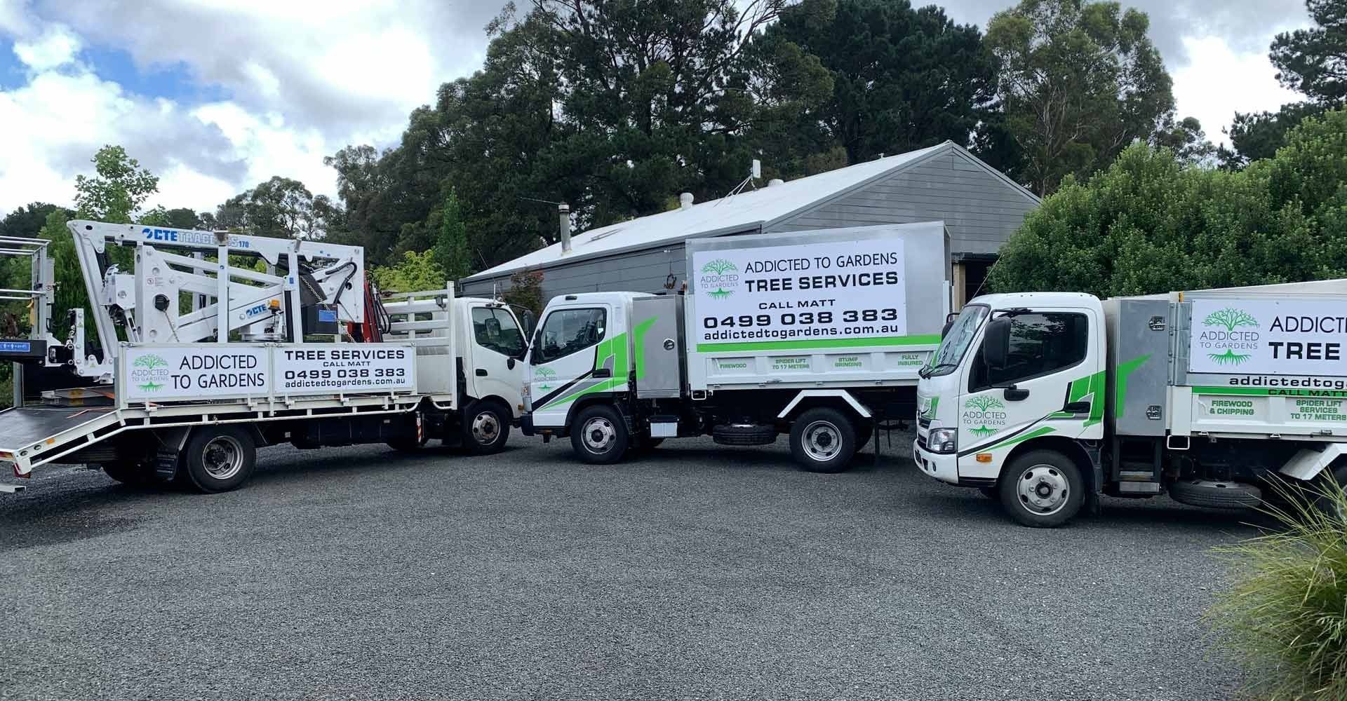 Four white and green service trucks parked on gravel, with a building and trees in the background.