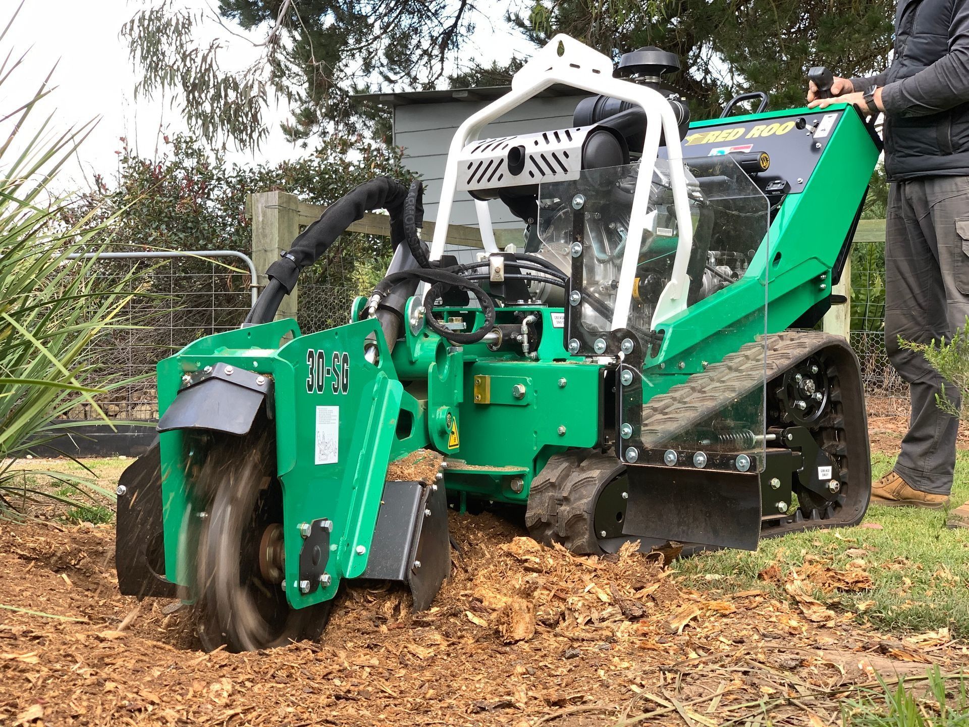 Green stump grinder on tracks grinding a tree stump; person operating it.