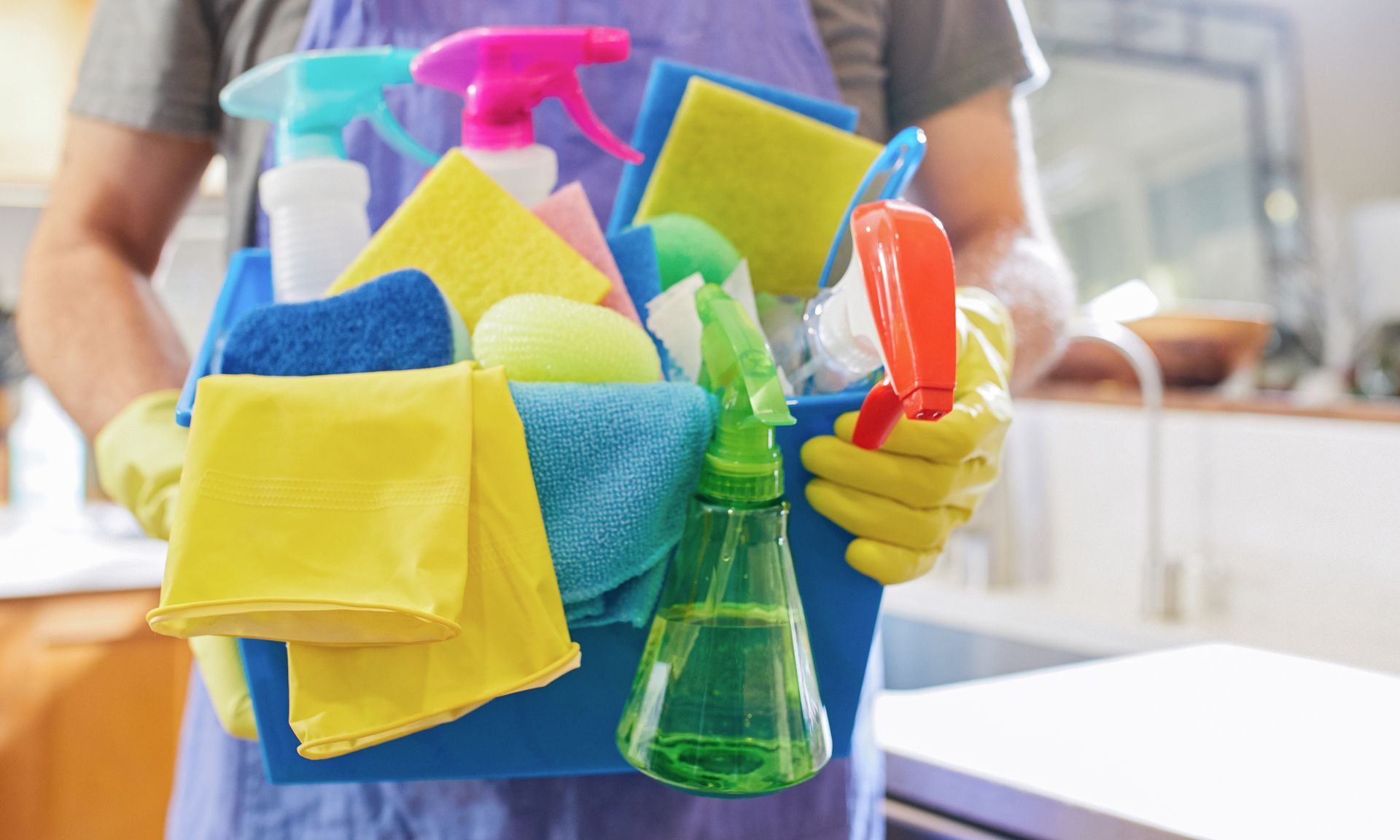 Person holding cleaning supplies in a blue bin. Yellow and blue cloths and spray bottles are visible.