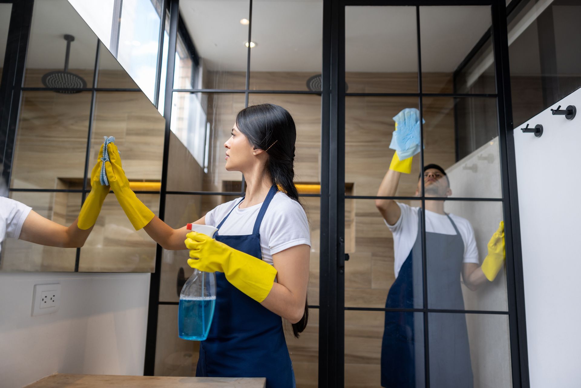 Woman and person in a bathroom cleaning glass with blue cleaning spray and yellow gloves.