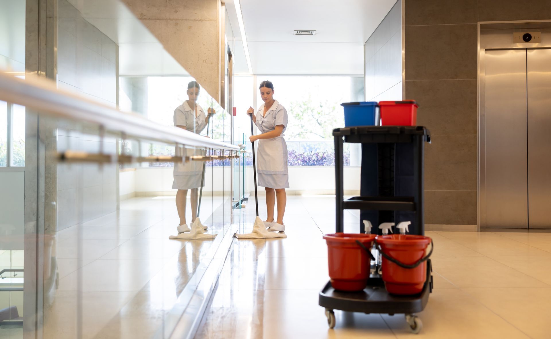 Woman mopping a shiny hospital hallway floor next to a cleaning cart.