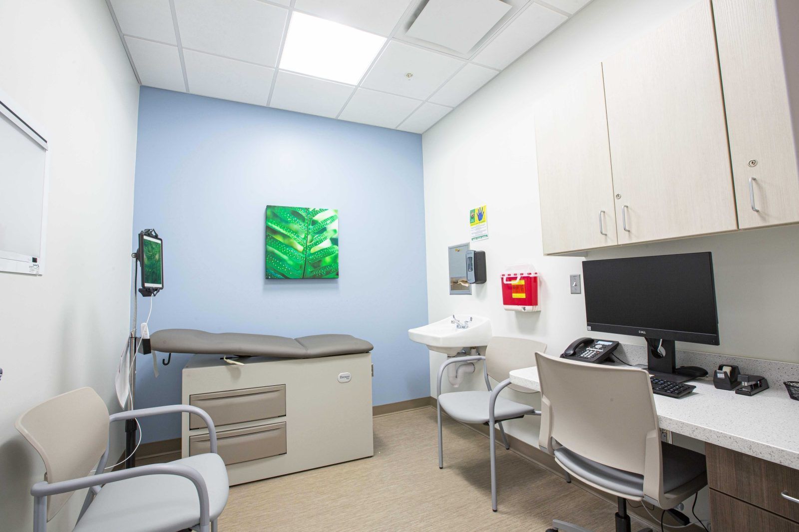 Medical exam room with exam table, sink, cabinets, desk, and computer in a clean white-and-blue clinic.