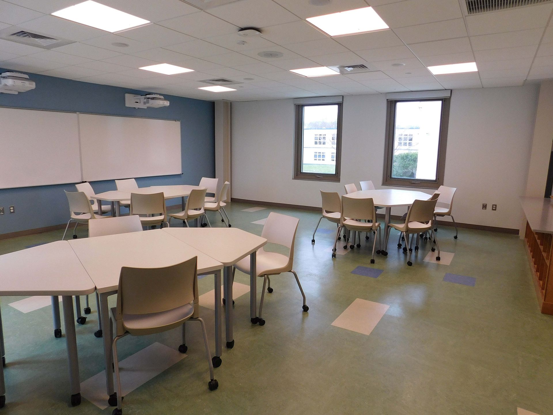 Empty classroom with tables, chairs, whiteboard, and two windows under fluorescent ceiling lights.