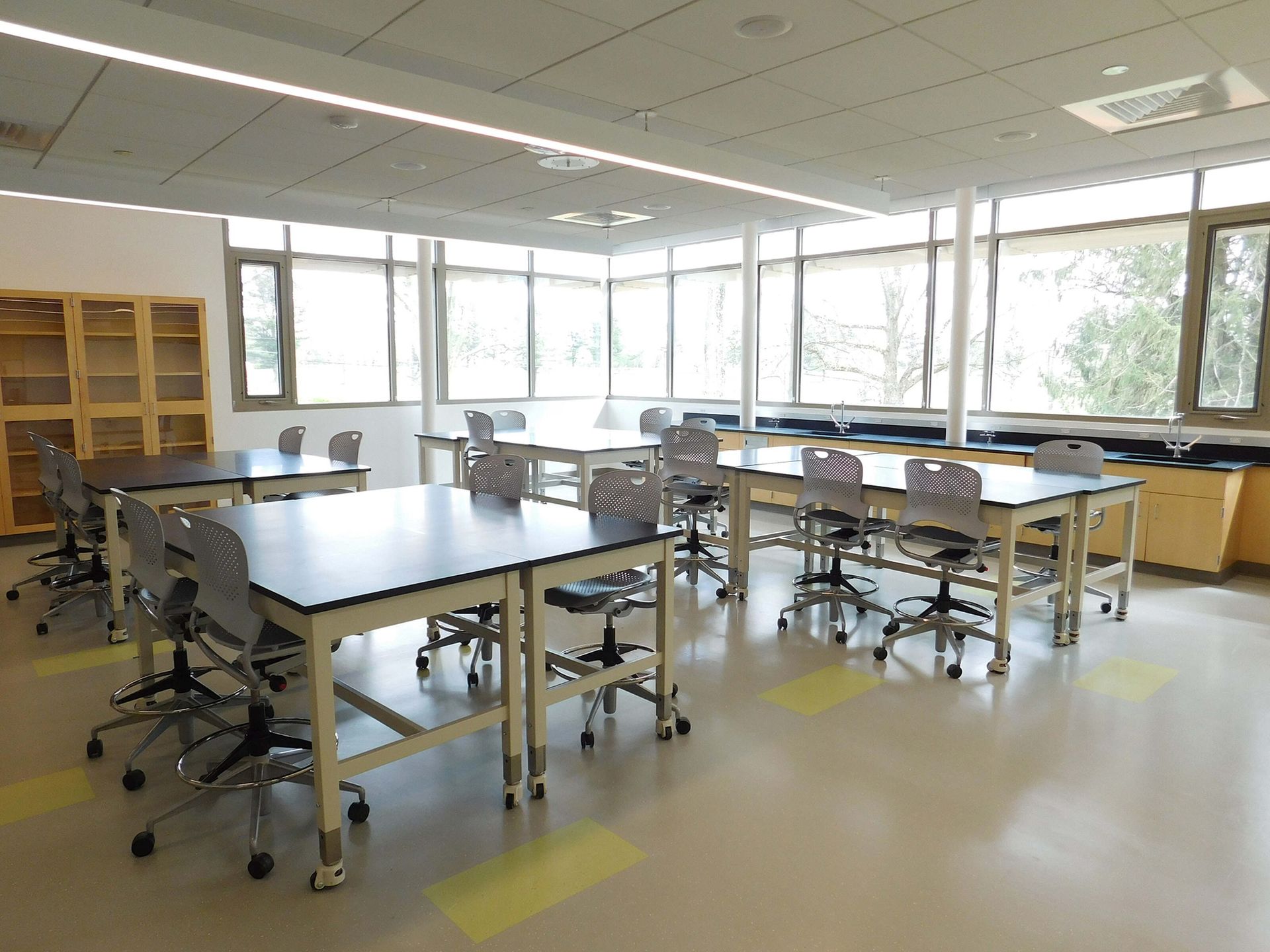 Empty science classroom with tables, stools, cabinets, and large windows letting in daylight