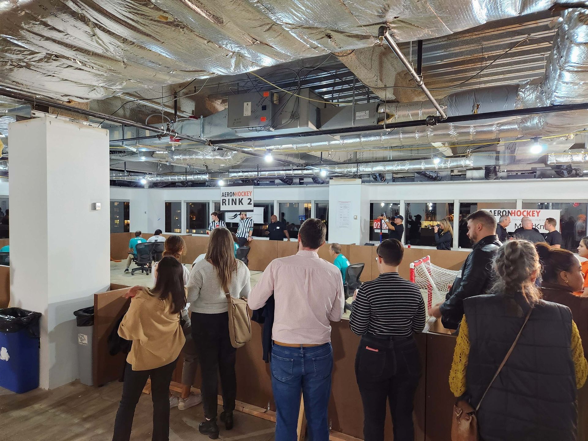 People gathered in a bright indoor space under exposed ceiling ductwork, facing a display area.