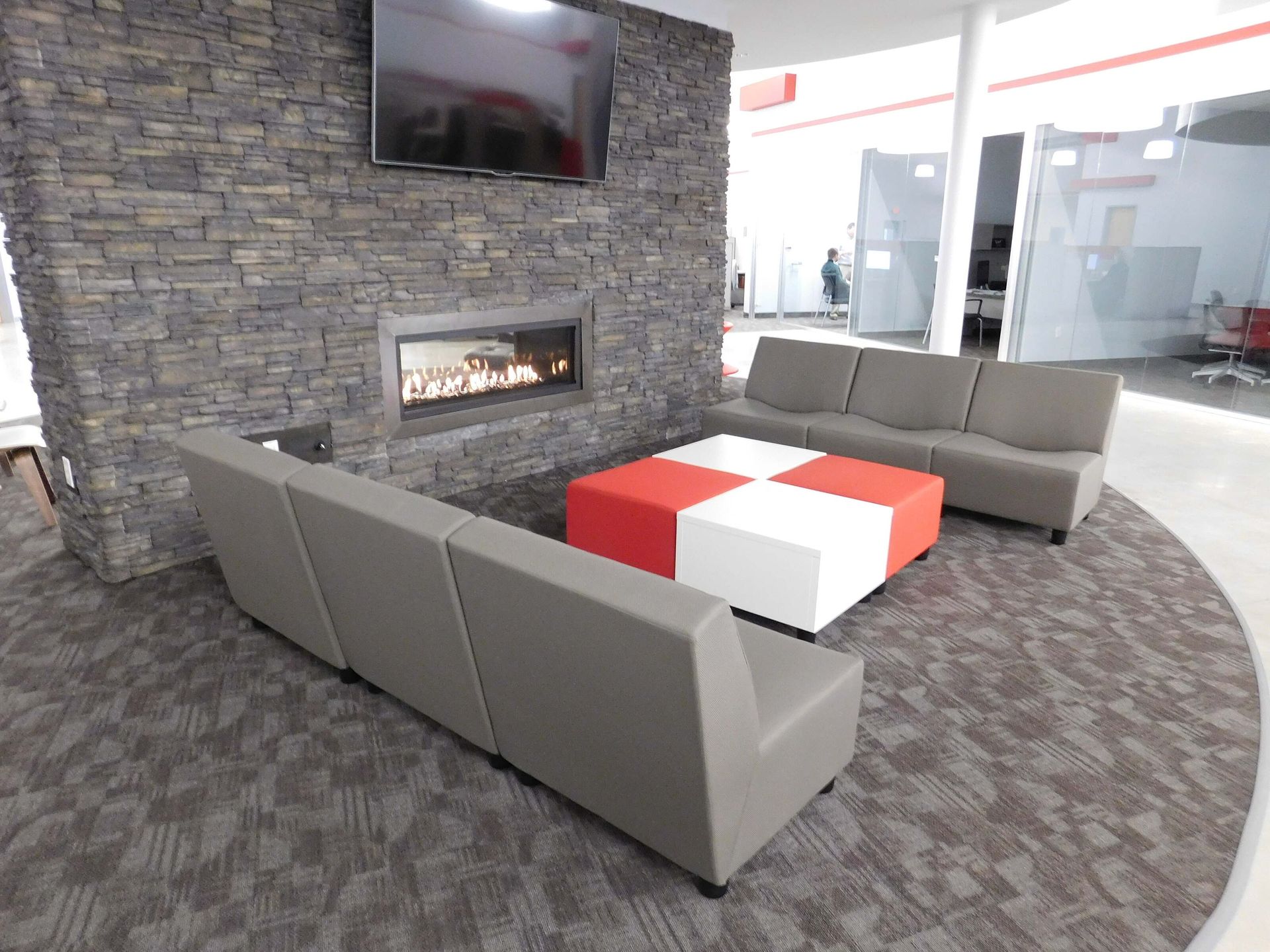 Modern lobby seating area with gray sofas, red-and-white ottoman, fireplace, and wall-mounted TV