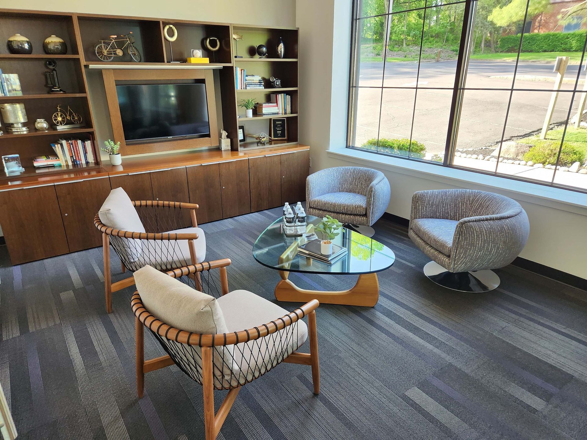 Modern lounge with wooden TV cabinet, round coffee table, two armchairs, and a window view of trees and road.