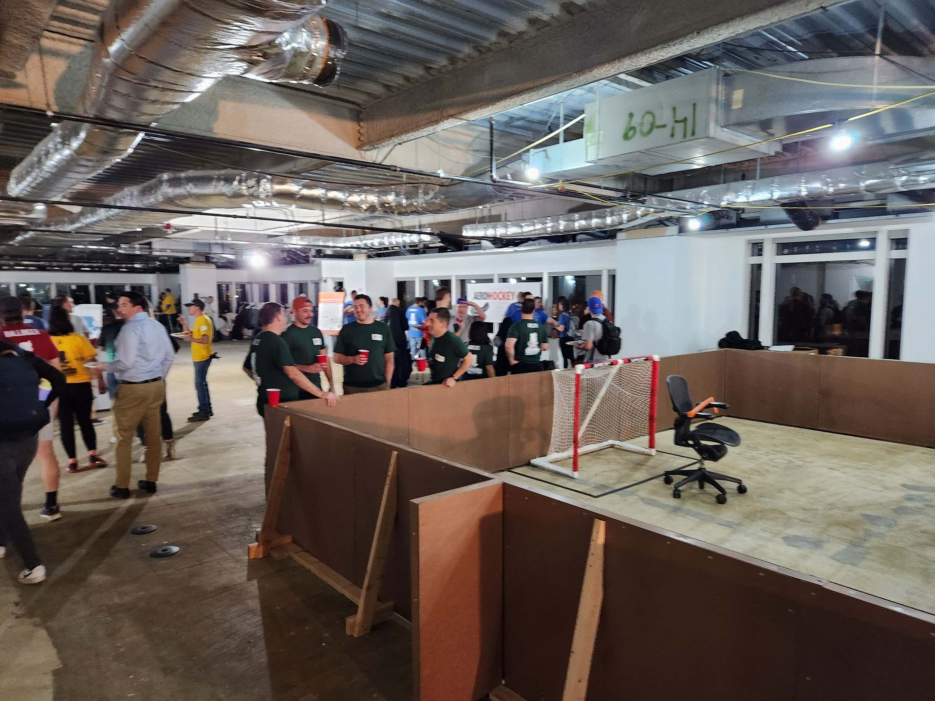 Indoor robotics event with people gathered around a red-and-white mini soccer field on a concrete floor.