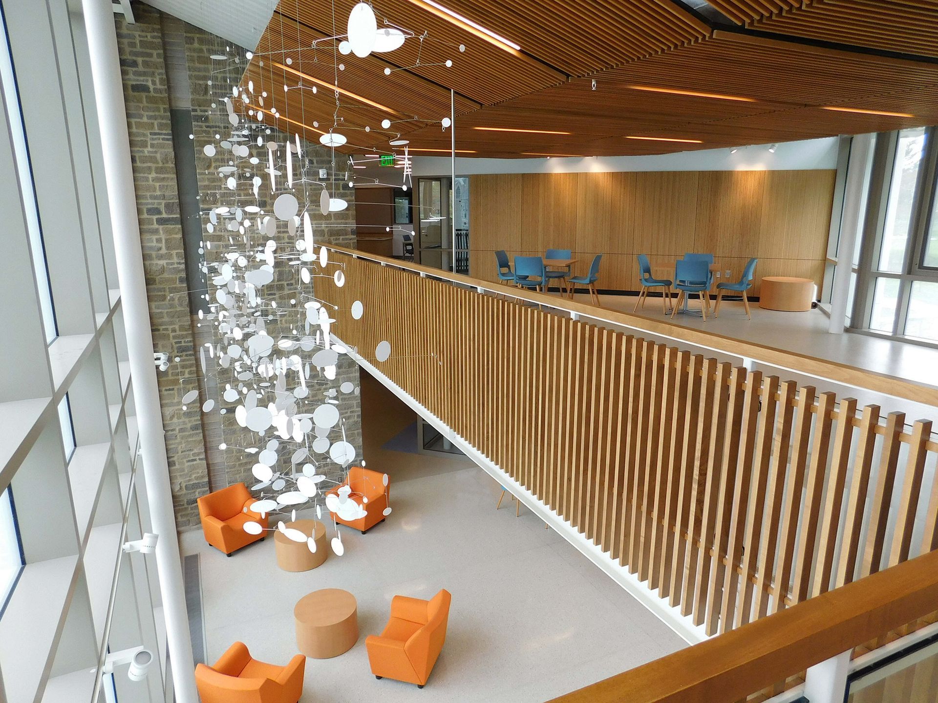 Modern atrium with hanging lights, orange chairs, and a wood slat railing overlooking a lounge area