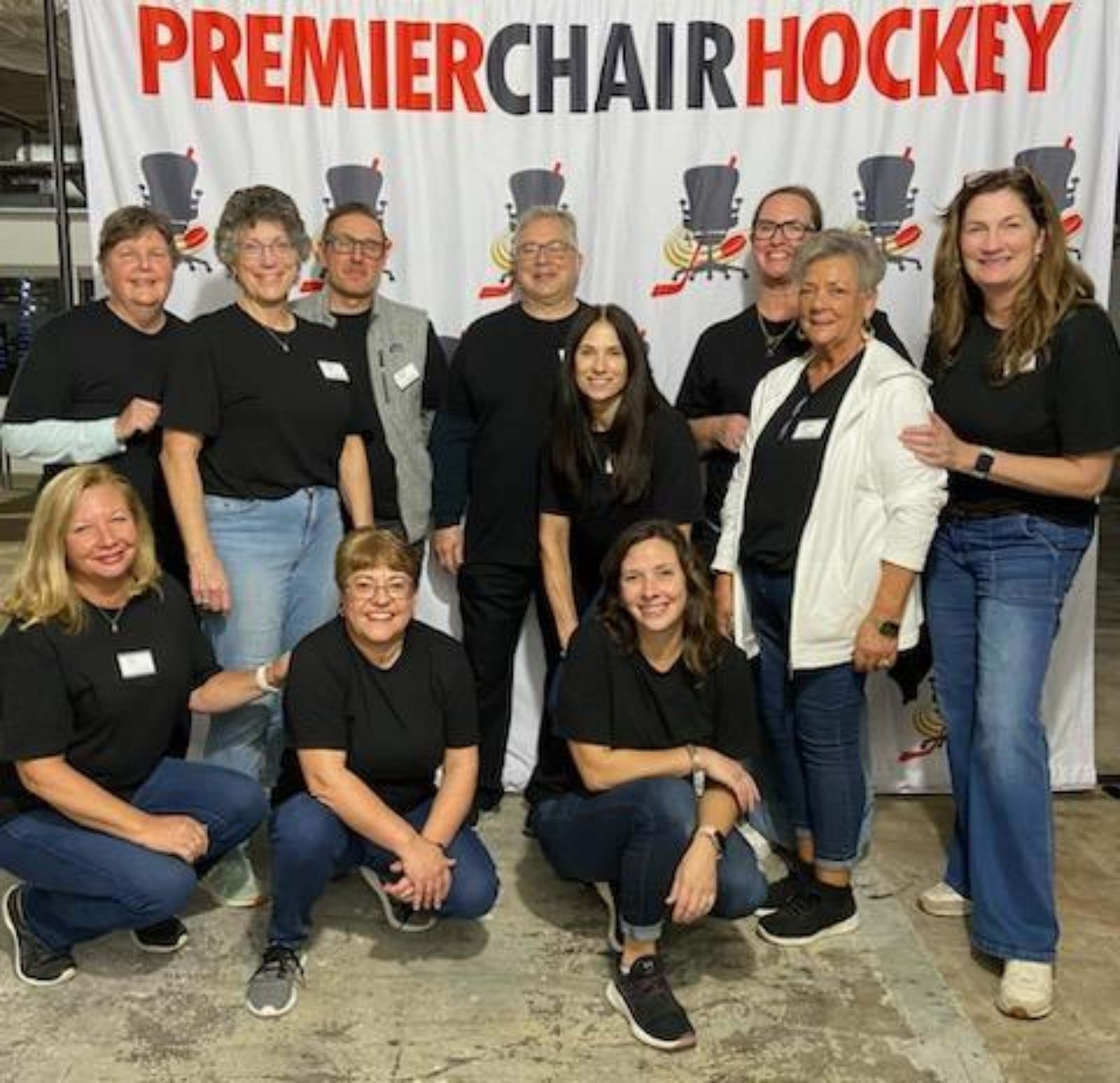 Team of people posing in front of a Premier Chair Hockey banner at an indoor event.
