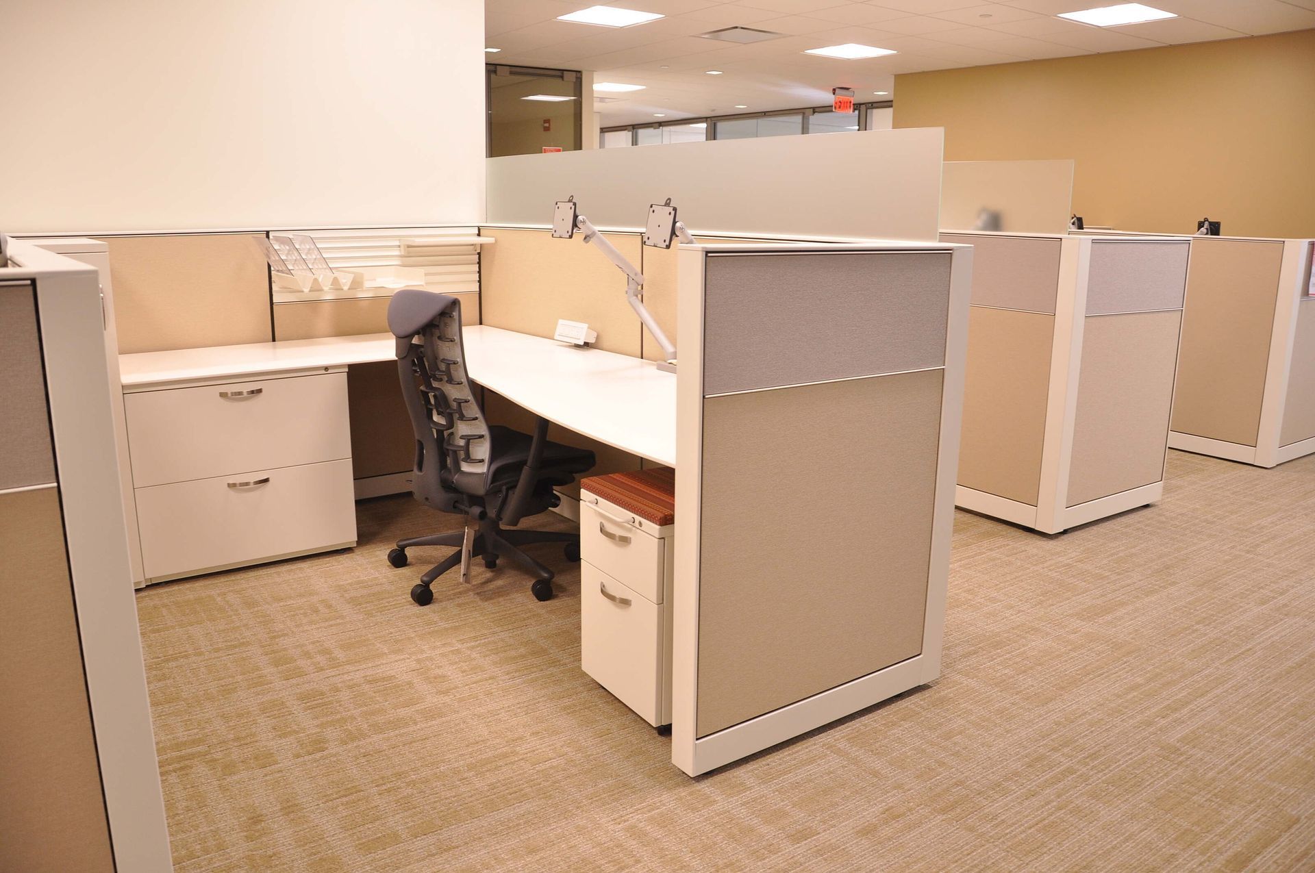Empty office cubicles with beige walls, a black chair, and carpeted flooring in an open workspace.