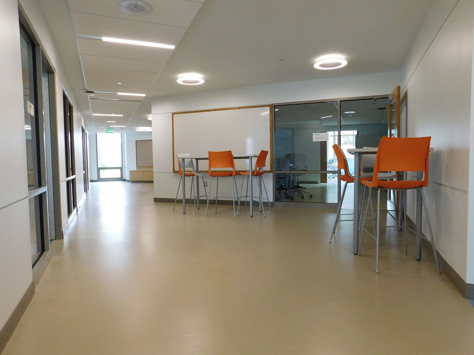 Modern hallway with orange chairs, tables, and glass-walled meeting area in the back