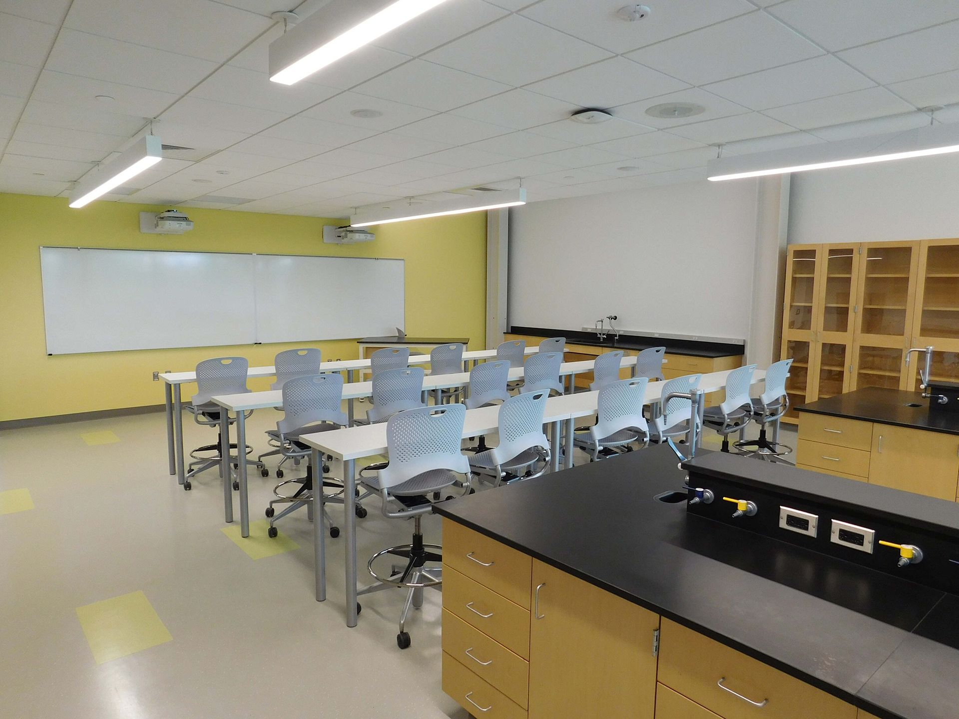 Bright science classroom with tables, stools, whiteboard, cabinets, and a lab counter under fluorescent lights
