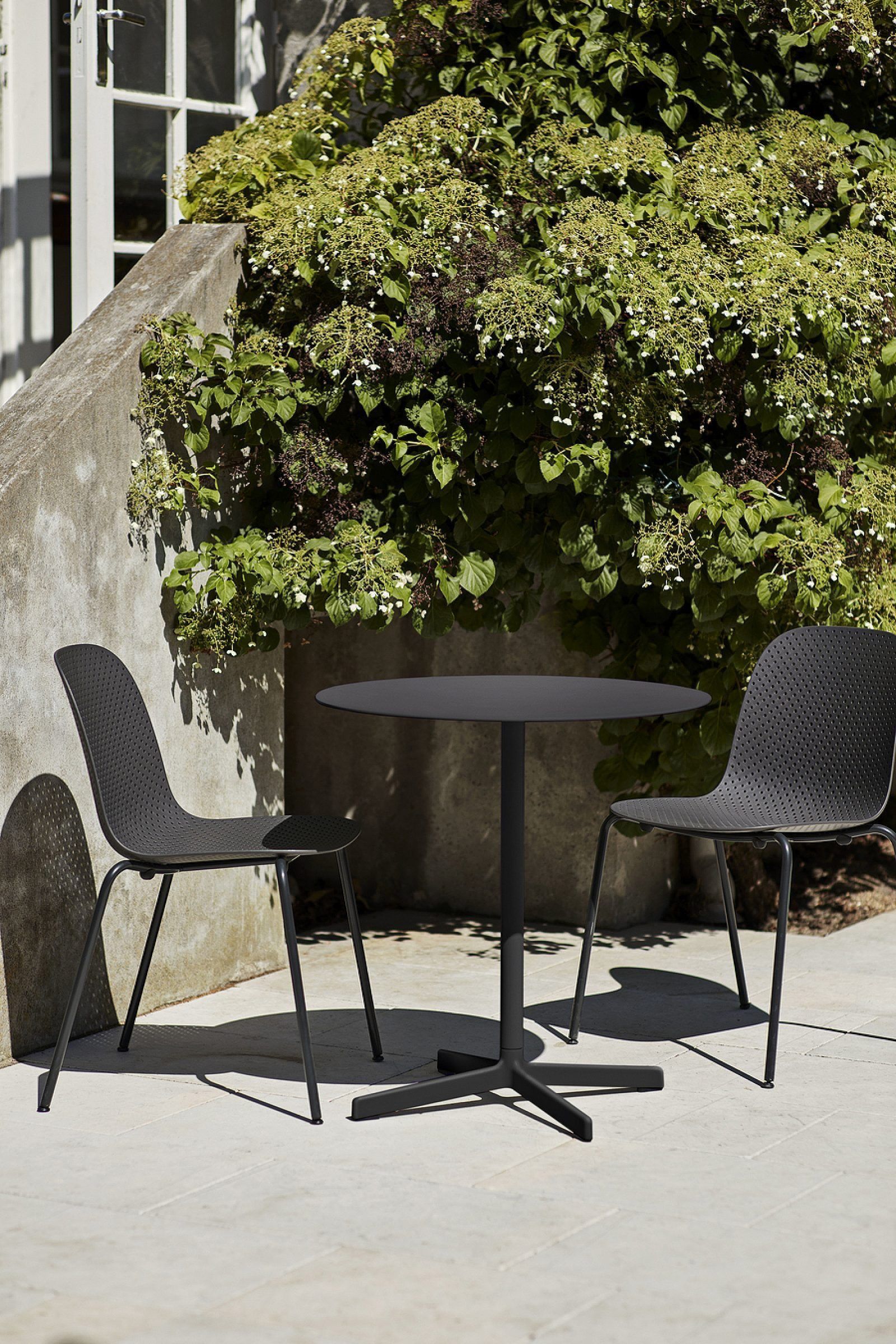 Two black patio chairs around a small round table on a sunlit concrete patio by a leafy hedge