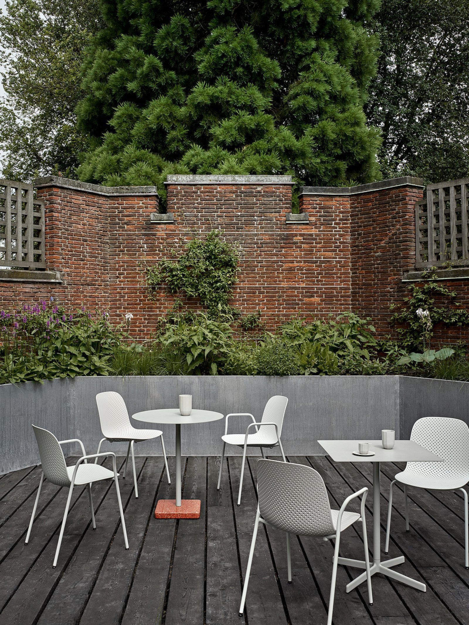 Two black patio chairs around a small round table on a sunlit concrete patio by a leafy hedge