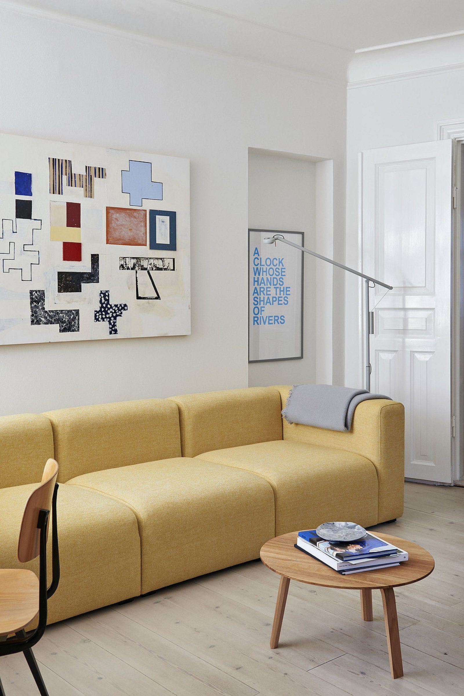 Bright living room with a yellow sofa, wooden coffee table, wall art, and a standing mirror near a white door.