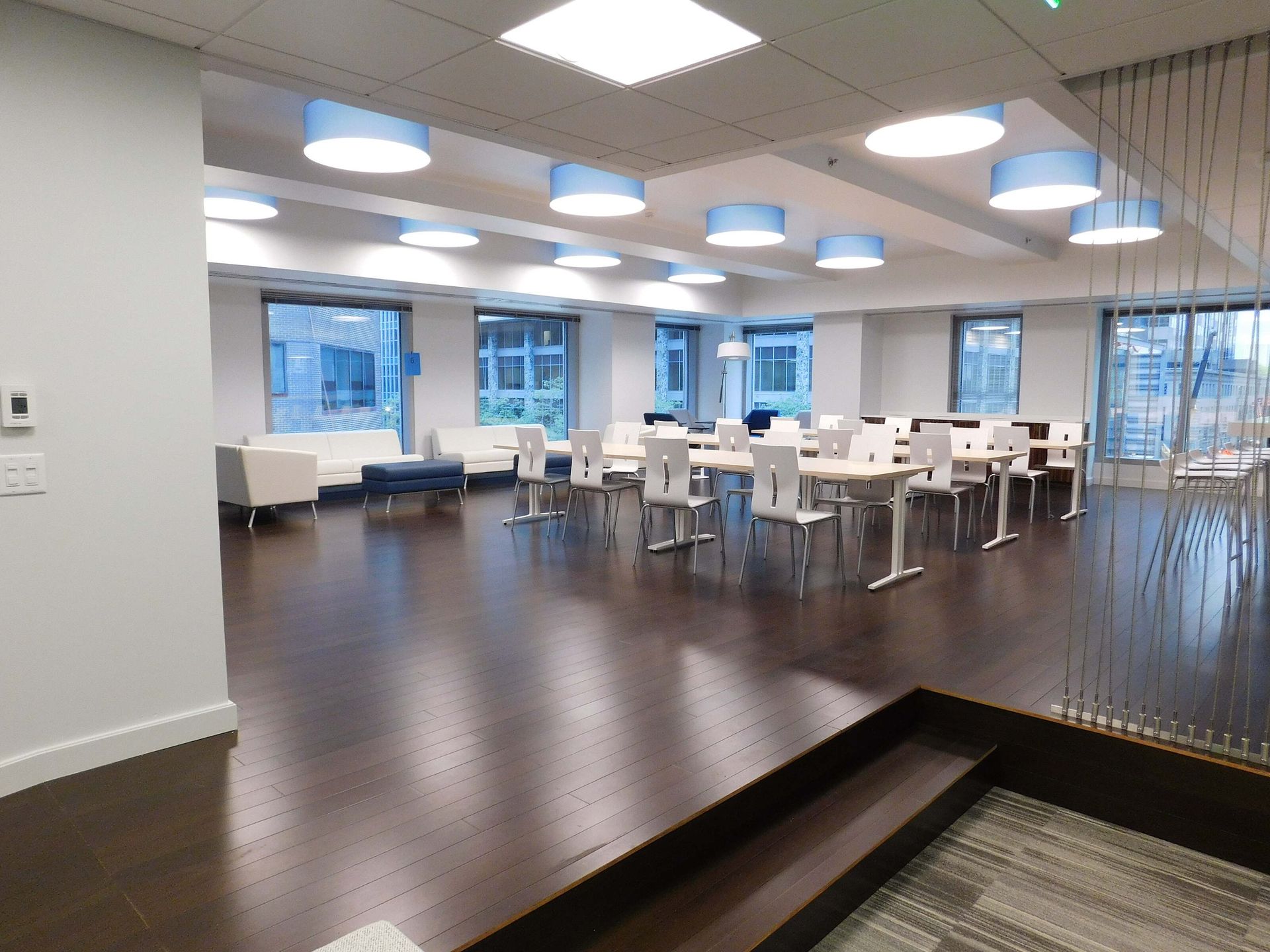 Bright modern office lounge with white tables, chairs, dark wood floor, and large windows.