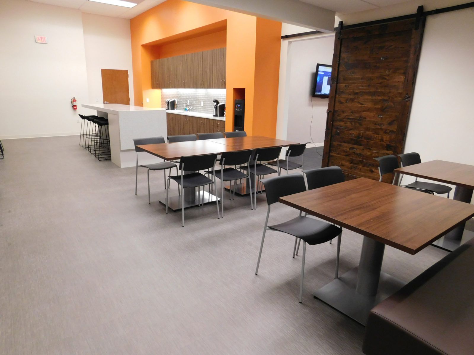 Empty modern cafeteria with gray carpet, wood tables, black chairs, and an orange service counter.