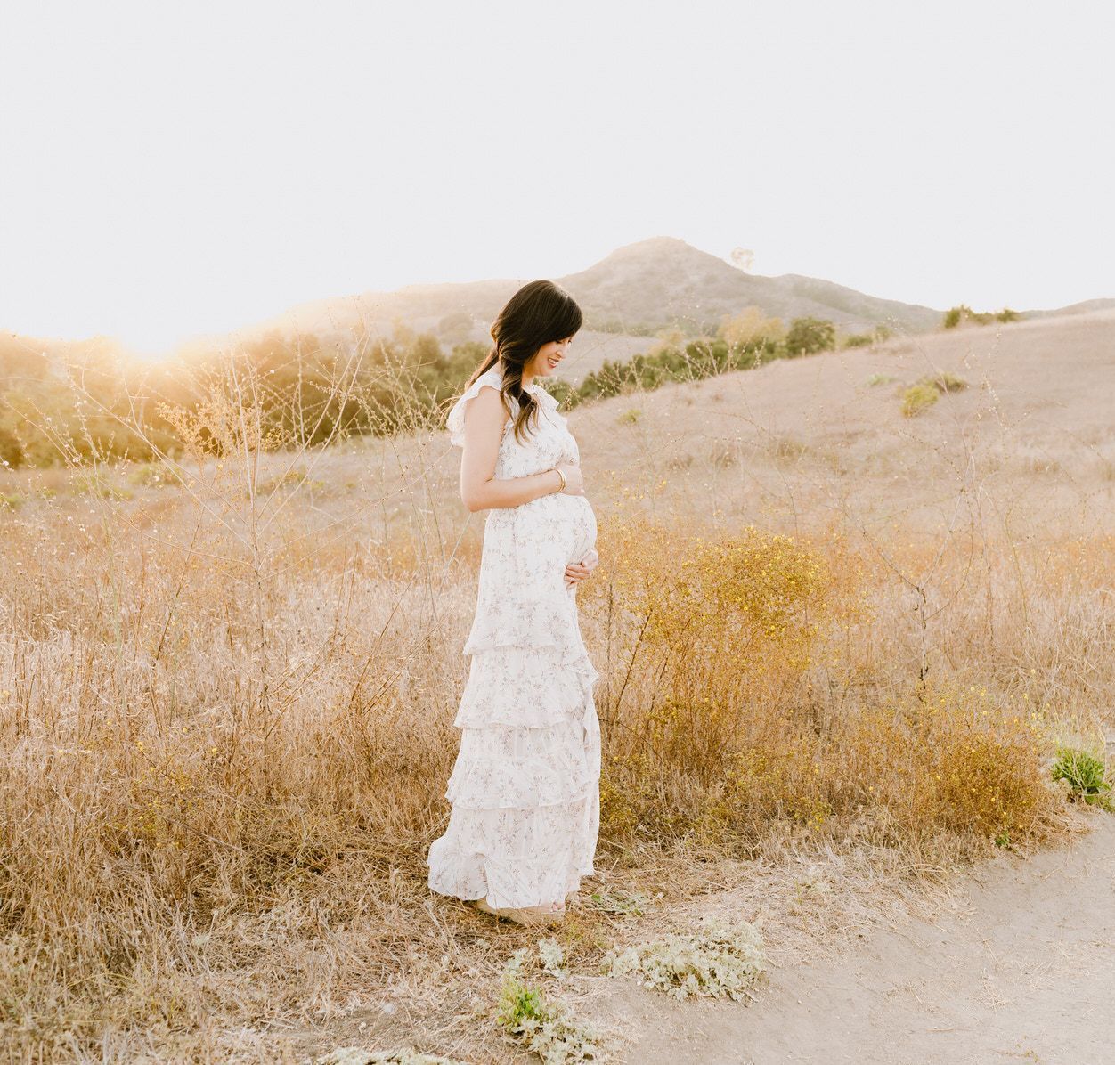 A pregnant woman in a white dress is standing in a field holding her belly.