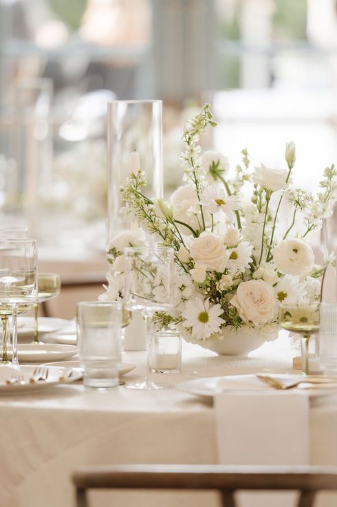 A photo with timeless editing of white floral centerpiece on a set dining table in a soft, bright room