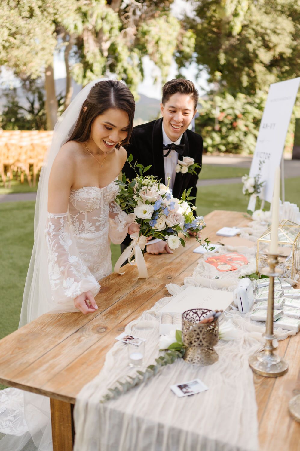 A bride and groom are signing their wedding invitations at a wooden table.