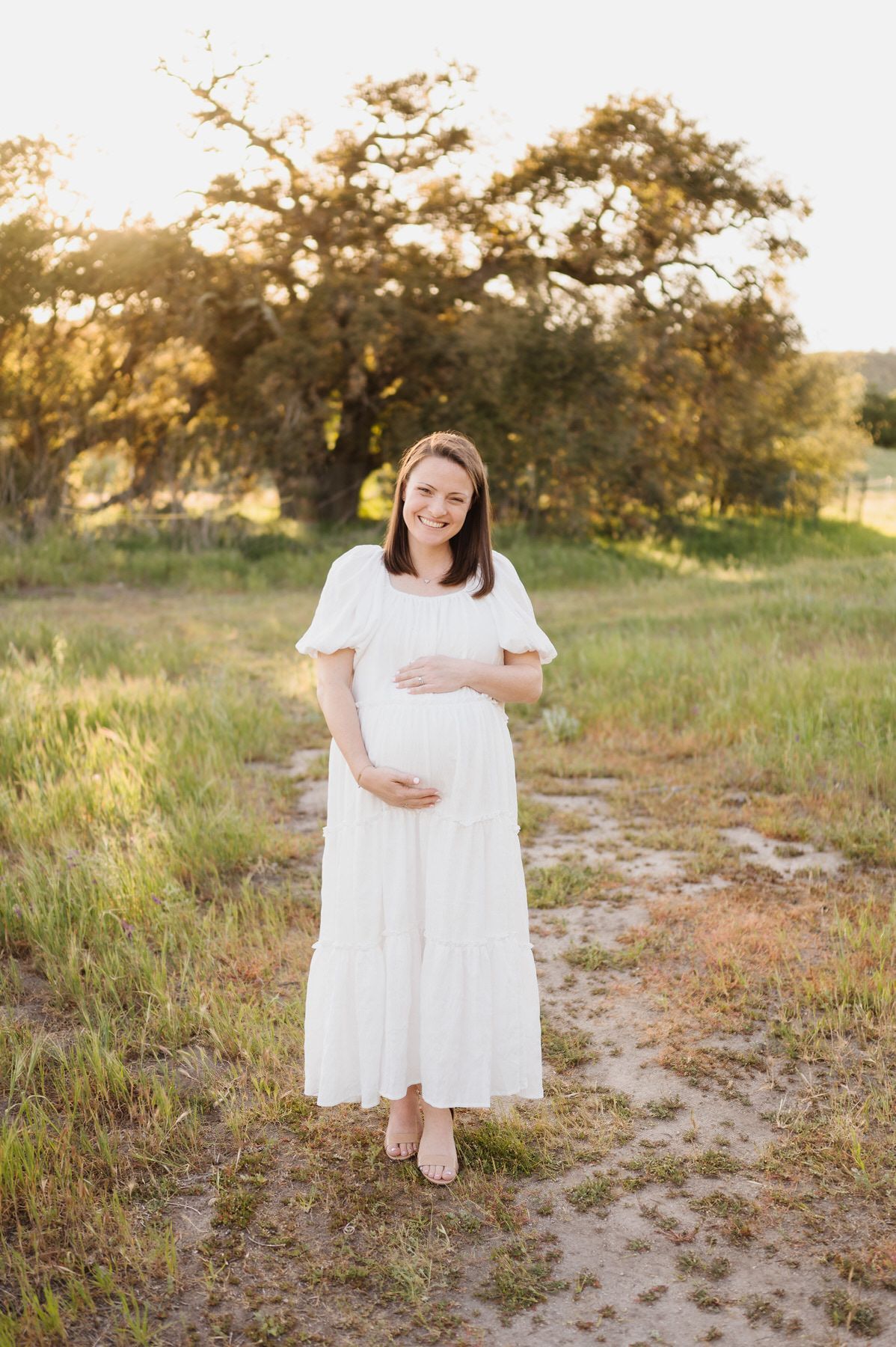A pregnant woman in a white dress is standing in a field holding her belly.