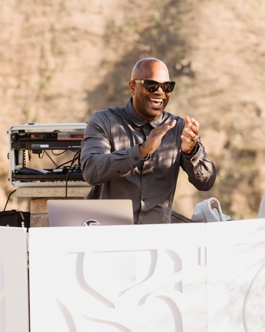 A man wearing sunglasses is standing behind a dj booth.