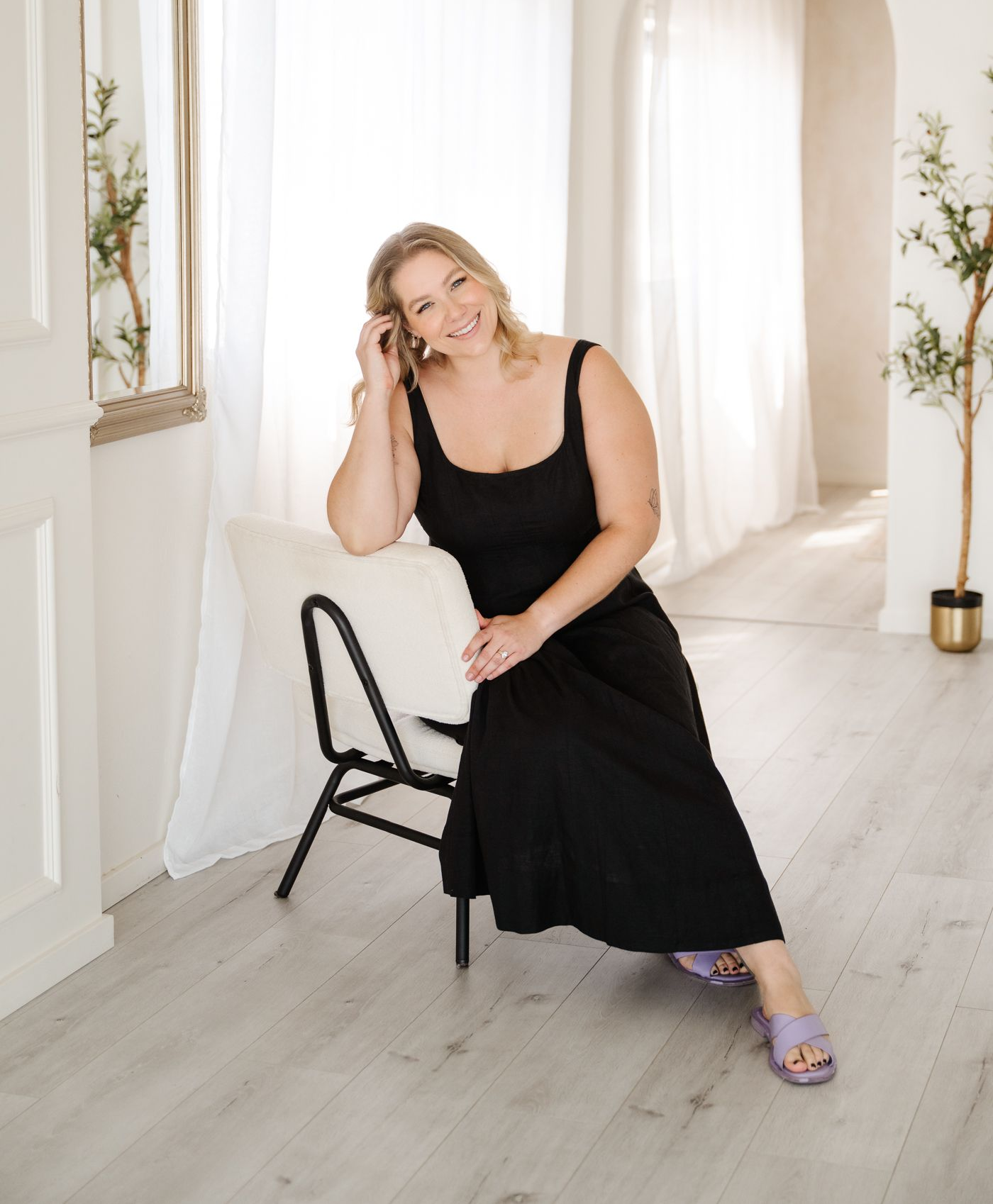 A person in a long black dress sits on a small black chair in a bright, modern room with white curtains and a mirror.