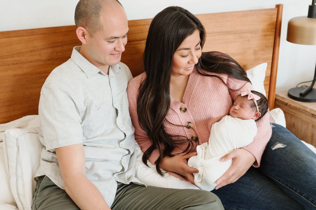 A man and woman are sitting on a bed holding a newborn baby for an in home newborn photo in Orange County.
