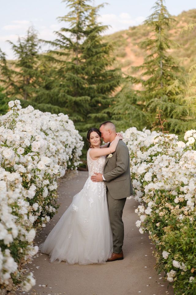 Bride and groom embracing on a flower-lined path in a garden with mountains in the background