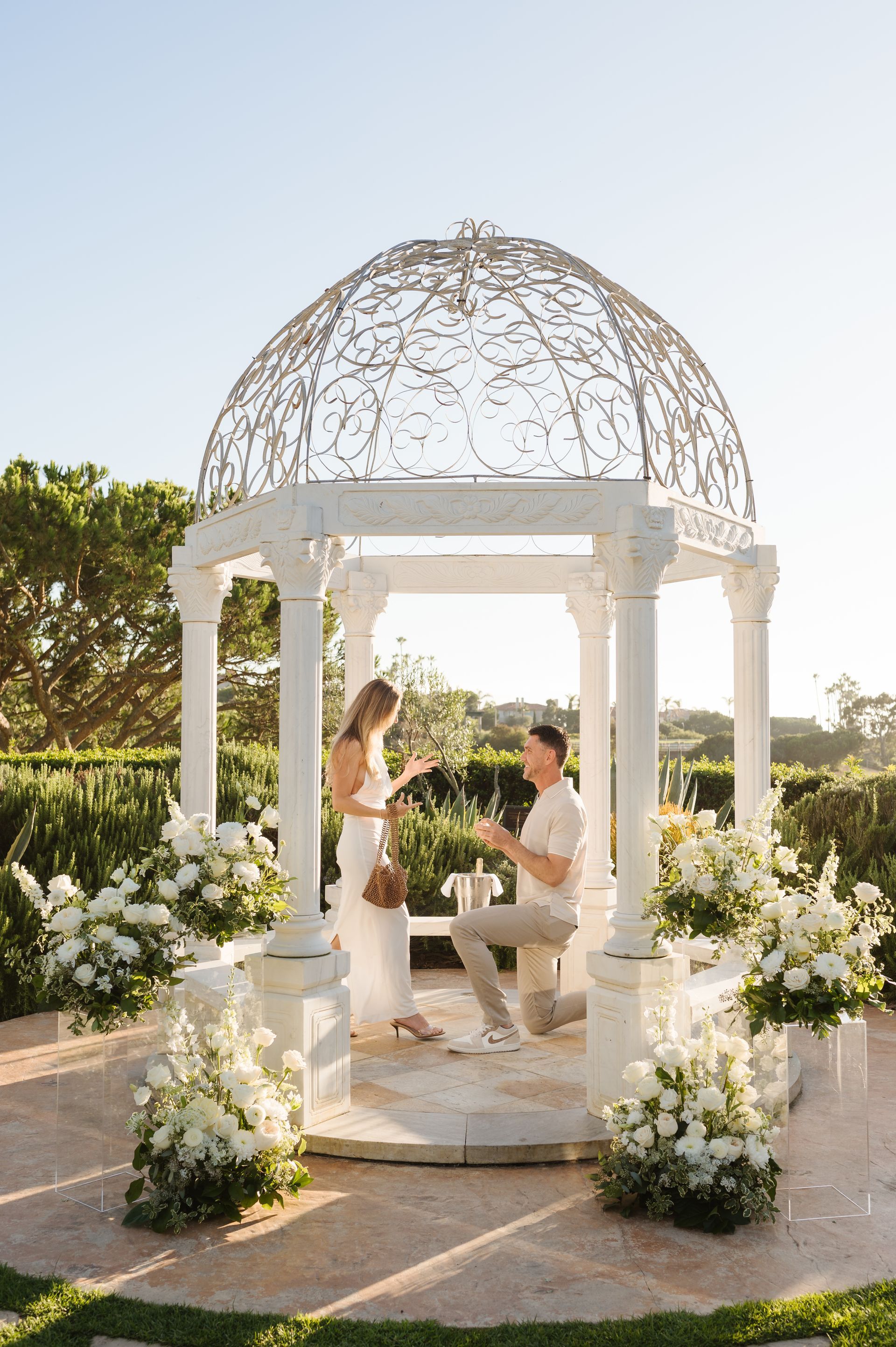 Man kneeling to propose in a white gazebo decorated with white flowers; woman reacts, sunny garden backdrop.