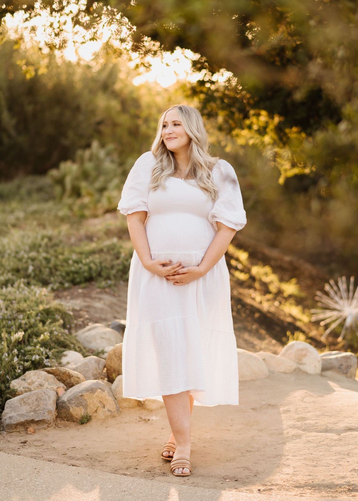 A pregnant woman in a white dress is standing in a park.