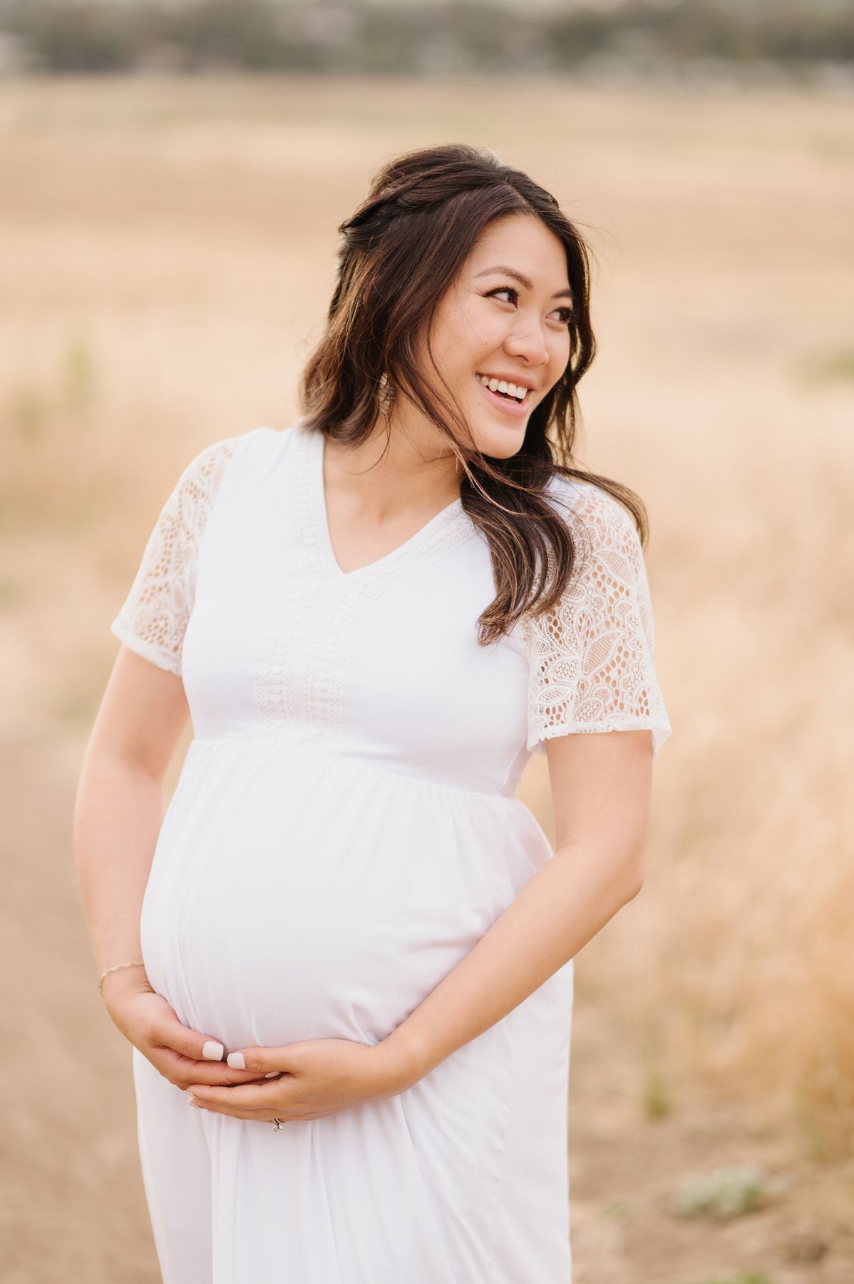 A pregnant woman in a white dress is standing in a field and smiling.