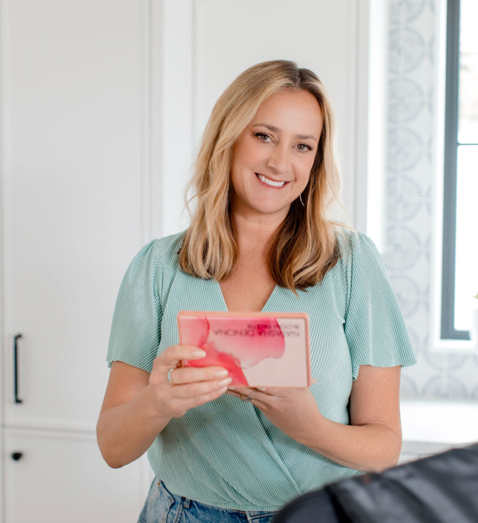 A person in a long black dress sits on a small black chair in a bright, modern room with white curtains and a mirror.