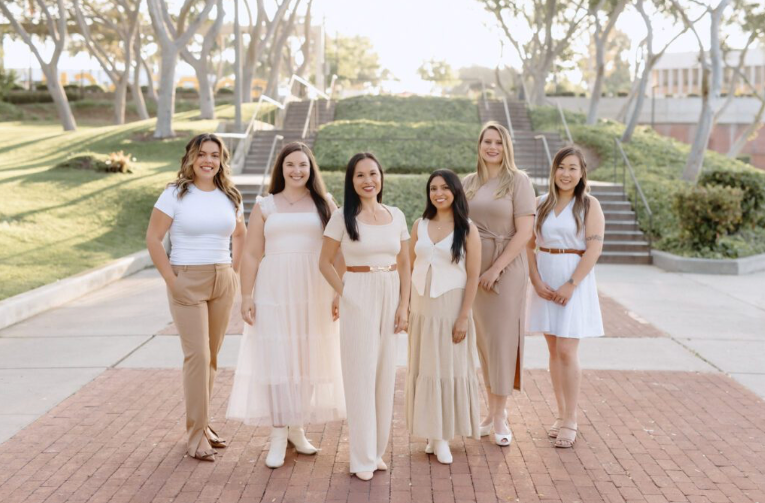 Six wedding planners in light-colored clothing stand together on a brick walkway outdoors with steps and trees in the background.