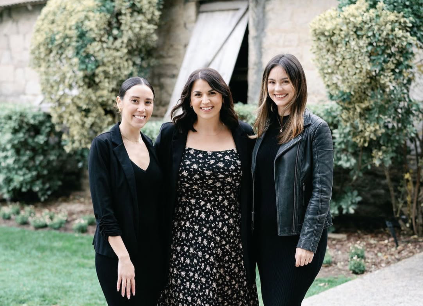 Three wedding planners stand side-by-side on a stone path in front of a building and greenery, all smiling while wearing black.
