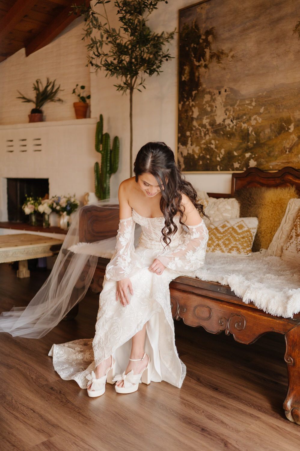 A bride in a wedding dress is sitting on a bench putting on her shoes.