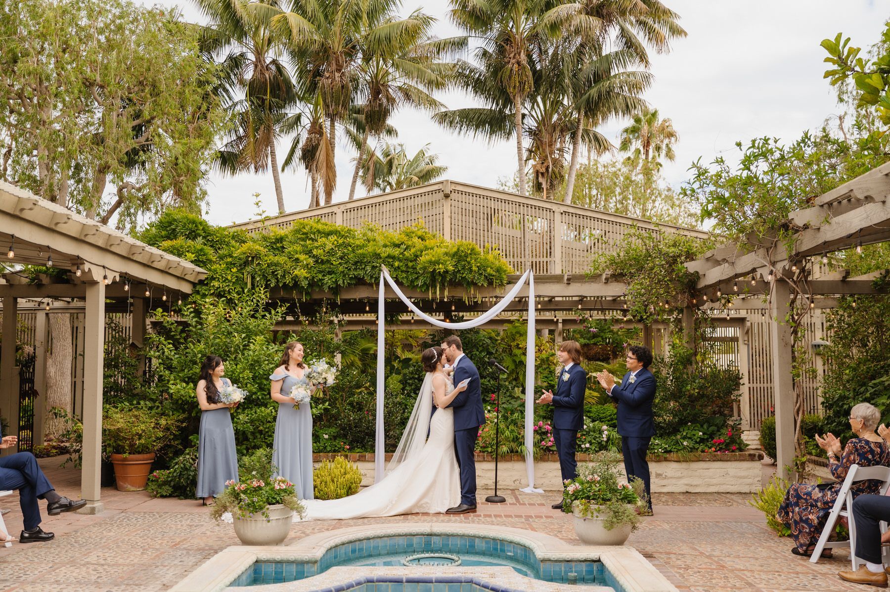 A bride and groom are kissing during their wedding ceremony in front of a pool.