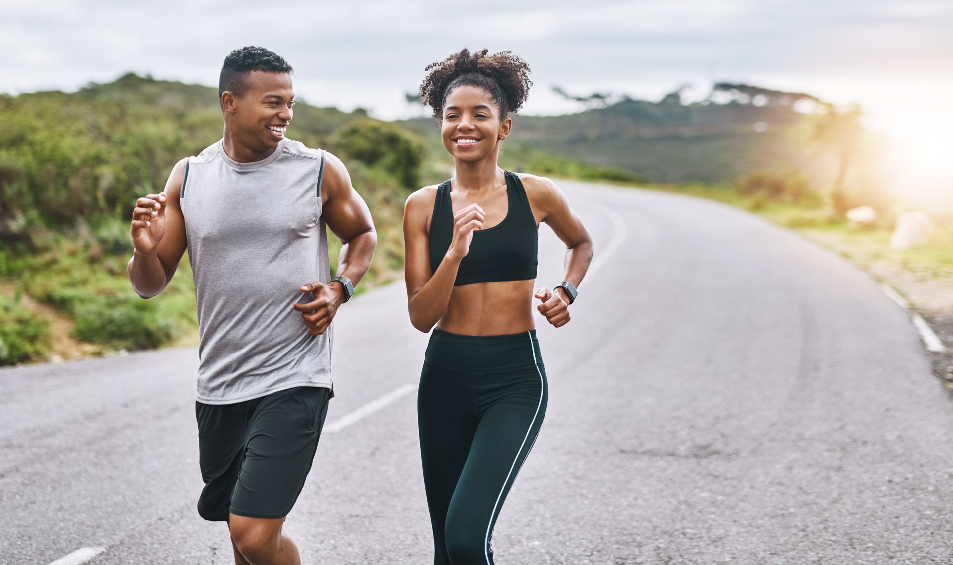 Couple running on a paved road; man in grey tank, black shorts; woman in black sports bra and leggings; sunny outdoors.