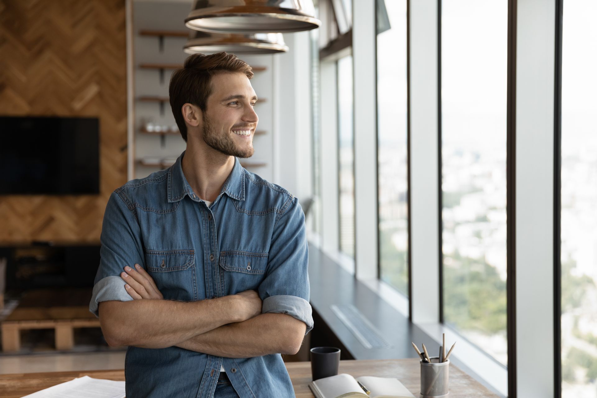 Man with arms crossed, smiling, looking out a window in a modern office setting.
