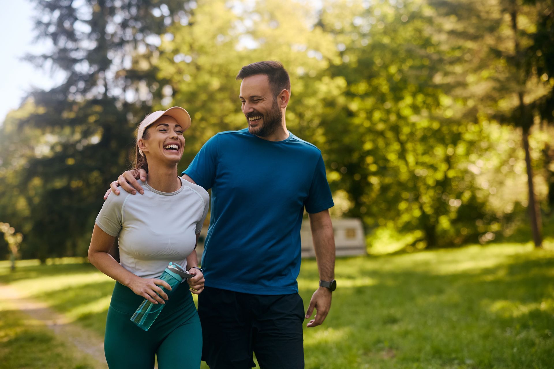A smiling couple in workout clothes walks arm-in-arm in a sunny park.