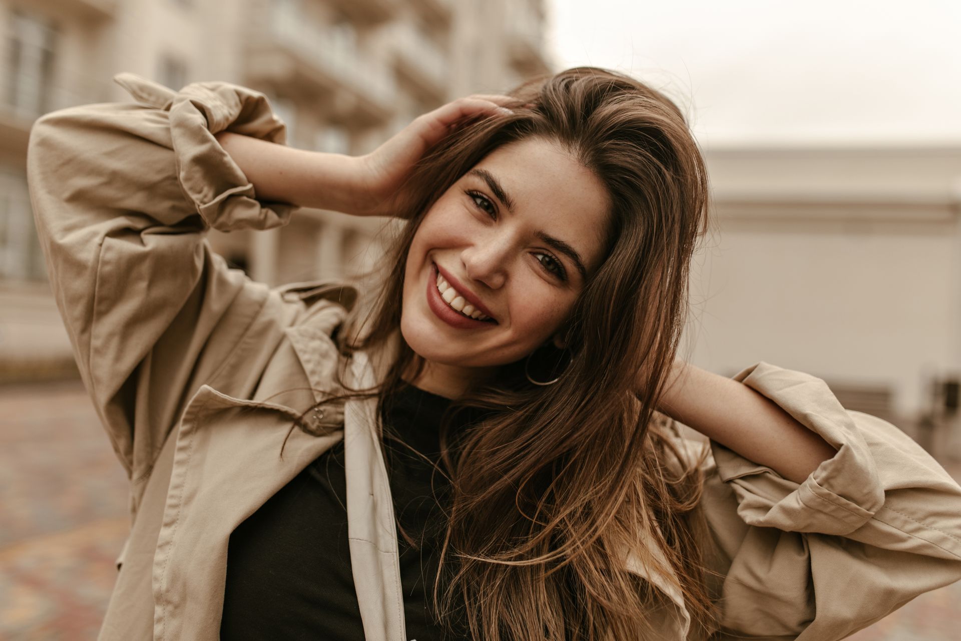 Woman with brown hair smiles, wearing a tan coat, hands in hair, outdoors.