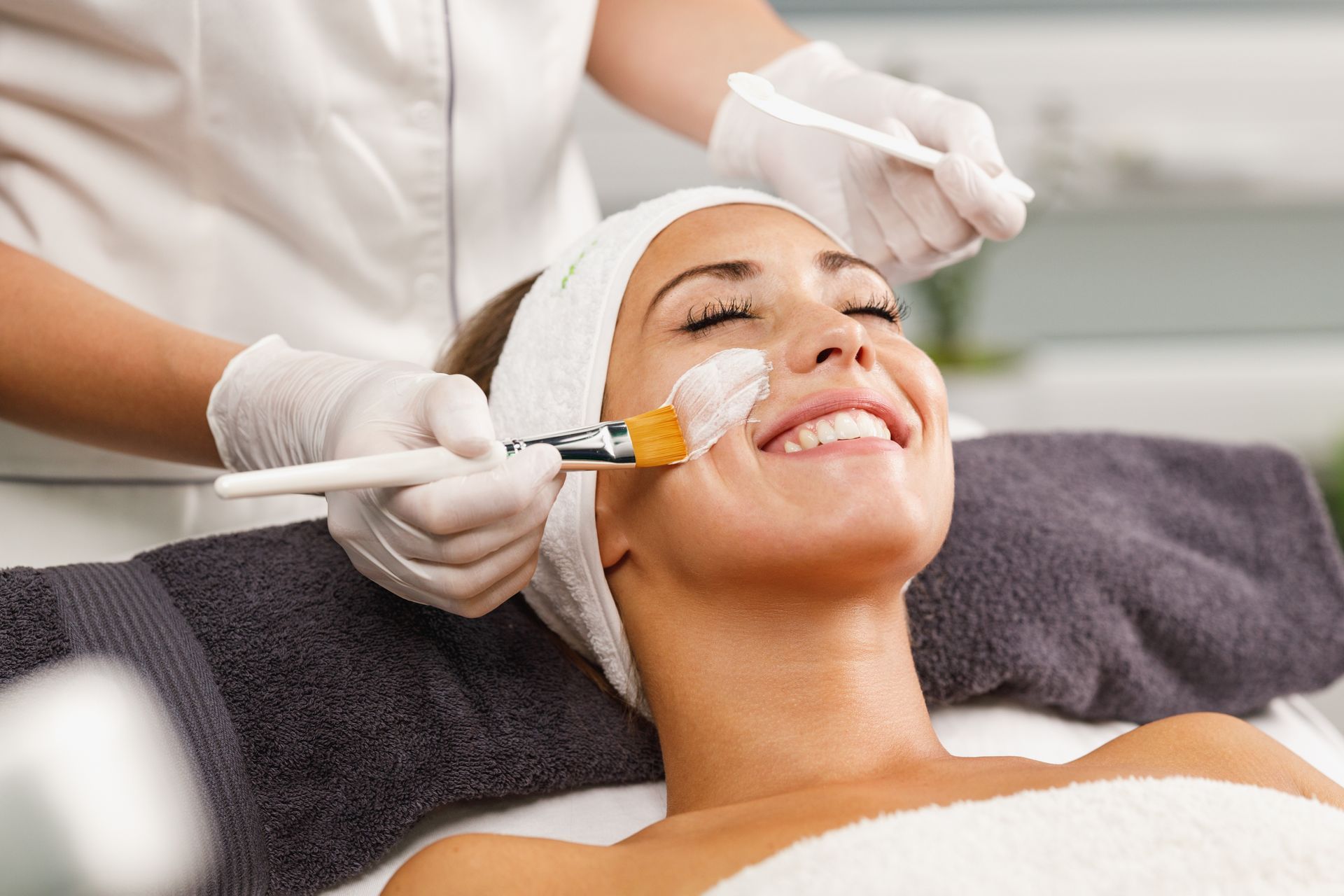 Woman receiving a facial treatment, smiling with eyes closed as esthetician applies a mask.