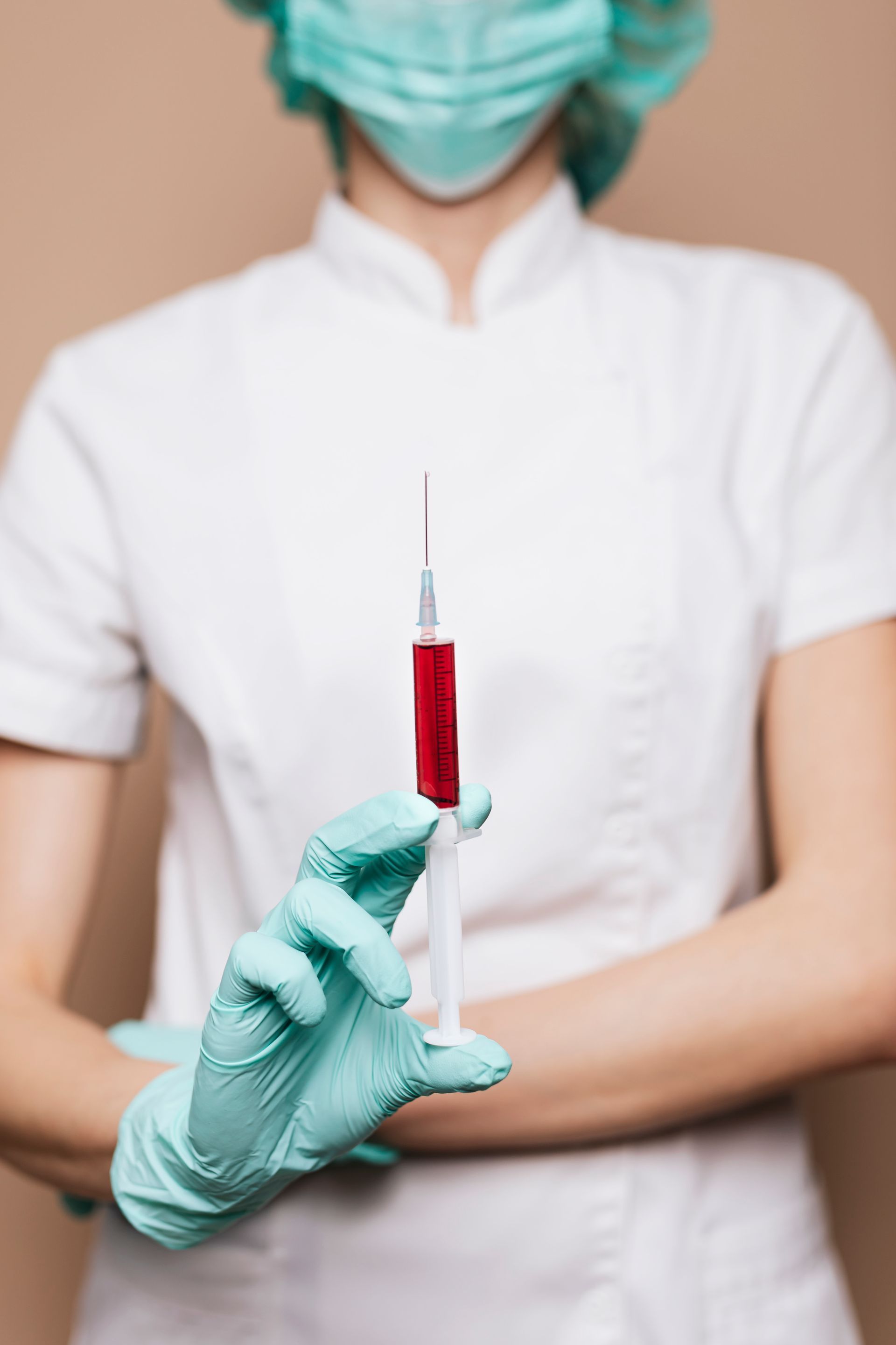 A nurse wearing a mask and gloves is holding a syringe with red liquid in it.