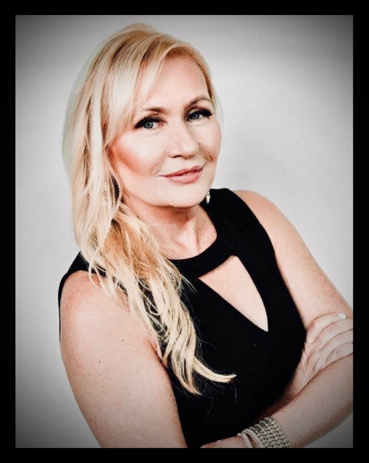Blonde woman in a sleeveless black top, smiling with arms crossed against a gray background