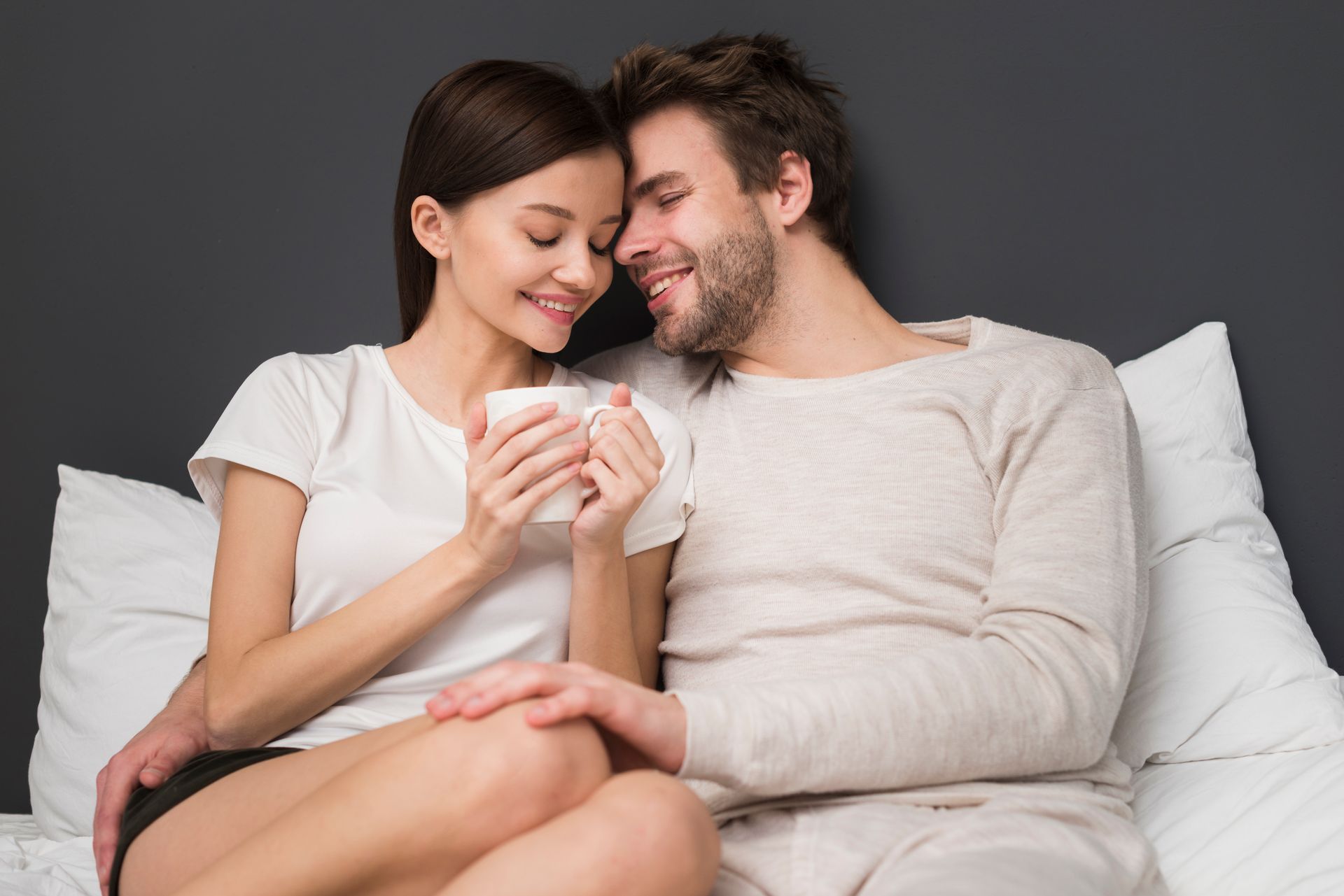 Couple in bed, smiling and holding a mug, cuddling on white pillows against a dark gray wall.