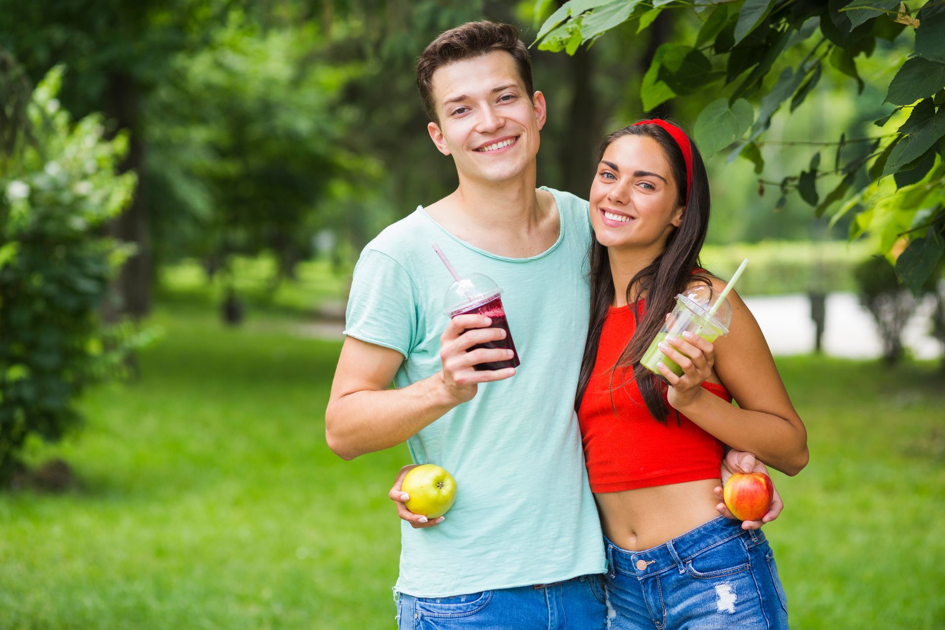 A man and a woman are standing next to each other in a park holding apples and drinks.