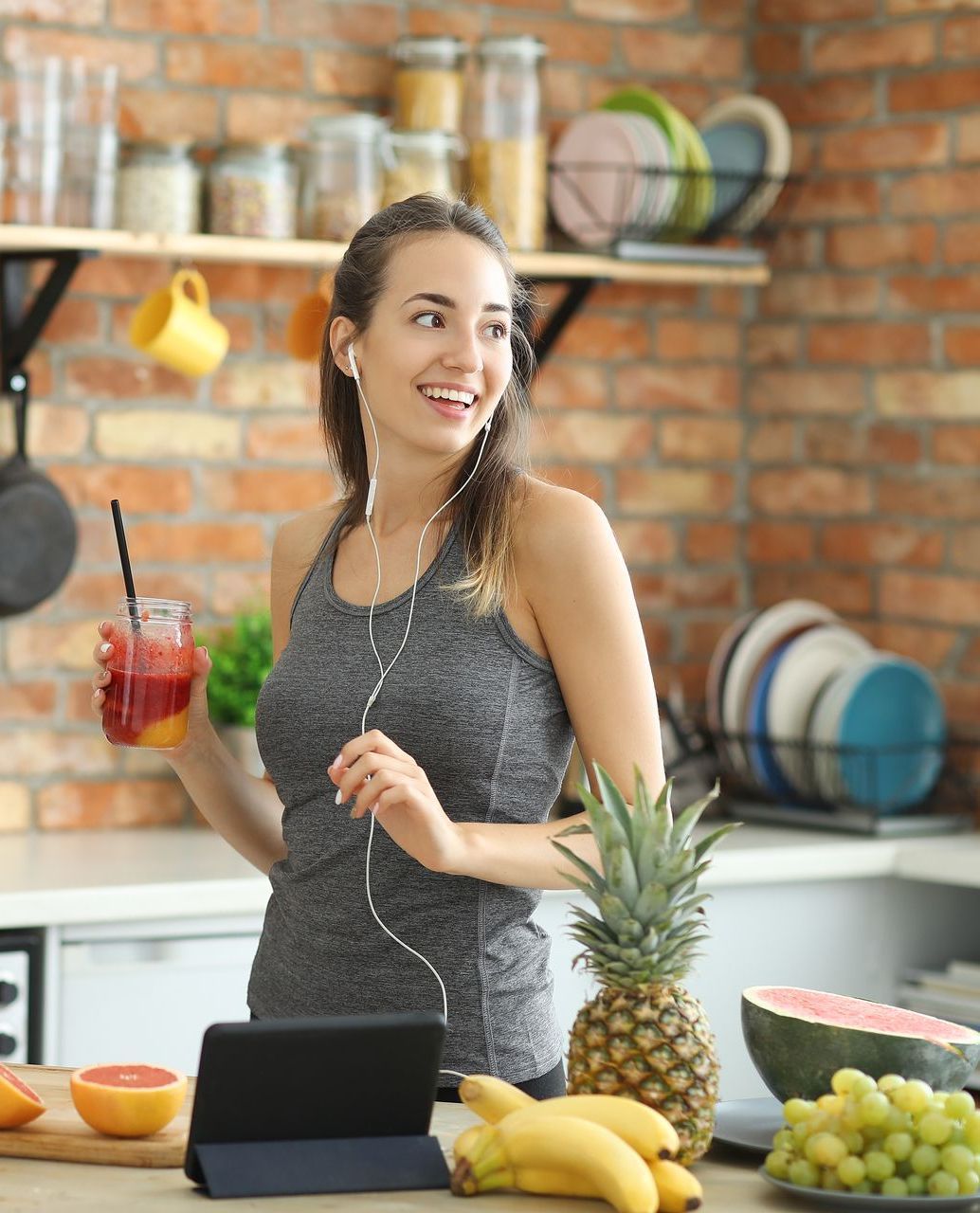 A woman is standing in a kitchen holding a drink and listening to music on a tablet.